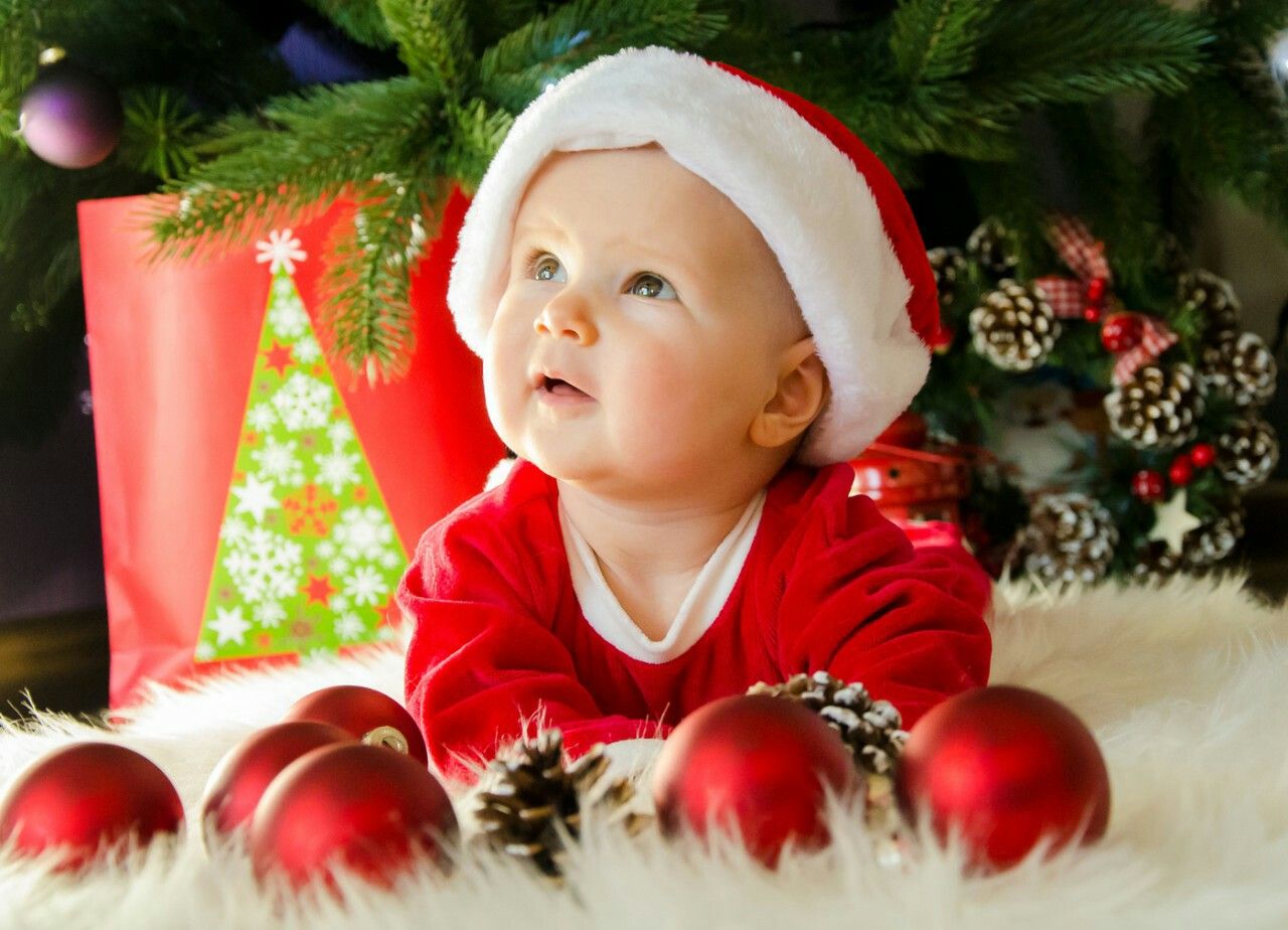 bébé habillé aux couleurs de Noël, couché au milieu de guirlandes et de boules de Noël, au pied d'un sapin décoré.